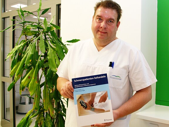 Pflegekraft in weißer Dienstkleidung hält ein Buch mit dem Titel „Schmerzpatienten behandeln“ und zeigt es in die Kamera. Das Cover zeigt eine medizinische Behandlung am Handrücken. Die Umgebung wirkt klinisch.
