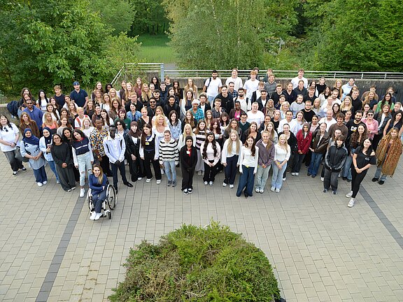 Großes Gruppenfoto vieler Menschen im Freien auf einem gepflasterten Platz, in mehreren Reihen angeordnet, zur Kamera gewandt; aufgenommen aus erhöhter Perspektive, im Hintergrund Bäume.