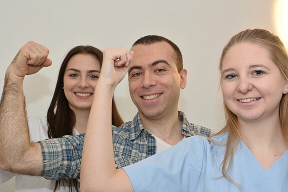 Drei Personen stehen nebeneinander und zeigen mit ihren Händen Muskeln. Alle lächeln freundlich in die Kamera. Die Person links trägt ein weißes Oberteil, die mittlere Person ein kariertes Hemd und die Person rechts ein hellblaues Krankenhausoberteil.