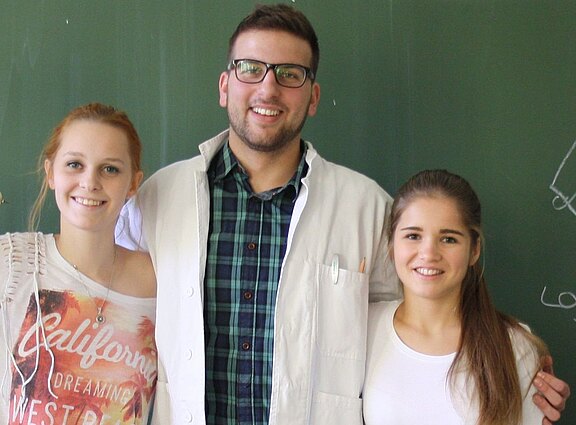 Vor einer Tafel stehen drei junge Menschen nah beieinander und lächeln. Die mittlere Person trägt Brille und weißen Laborkittel, die beiden anderen Freizeitkleidung. Sie legen einander die Arme um die Schultern, wirken freundlich und verbunden.