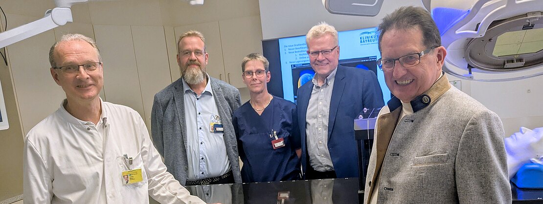 Fünf Erwachsene stehen in einem Klinikraum um ein großes Medizingerät. Links ein Arzt im weißen Kittel, daneben Mitarbeitende in beruflicher Kleidung. Im Hintergrund Bildschirme und Technik. Eindruck: Präsentation oder Einweisung in neue Ausstattung.