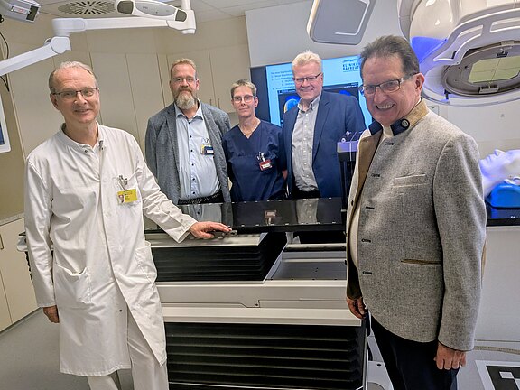 Fünf Erwachsene stehen in einem Klinikraum um ein großes Medizingerät. Links ein Arzt im weißen Kittel, daneben Mitarbeitende in beruflicher Kleidung. Im Hintergrund Bildschirme und Technik. Eindruck: Präsentation oder Einweisung in neue Ausstattung.