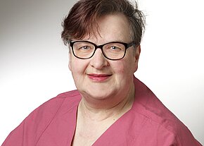 Porträt einer lächelnden Person mit Kurzhaarfrisur und schwarzer Brille vor hellem Hintergrund. Sie trägt ein rosa Oberteil, ähnlich einer Arbeitskleidung. Der Fokus liegt auf dem freundlichen Gesichtsausdruck.