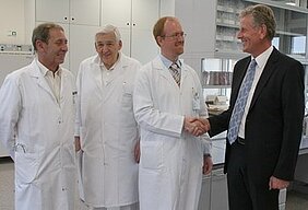Vier Männer in weißen Laborkitteln stehen in einem modernen Labor. Zwei von ihnen schütteln sich die Hände, während die anderen zuschauen. Die Atmosphäre wirkt freundlich und kollegial.