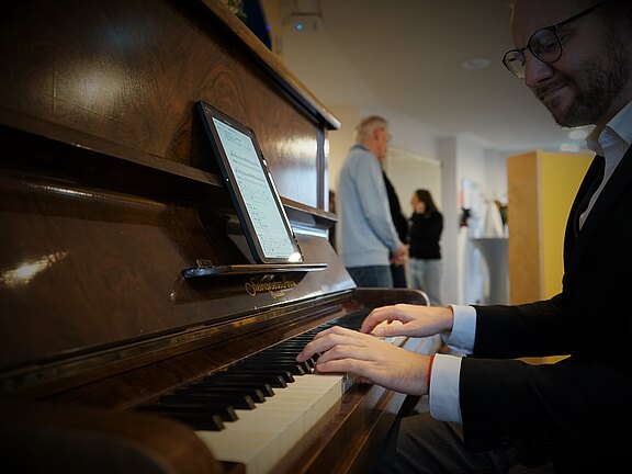 Nahaufnahme einer Person im Anzug, die ein aufrechtes Klavier spielt. Ein Tablet auf dem Notenpult zeigt digitale Noten. Fokus auf den Händen auf den Tasten, Hintergrund unscharf.