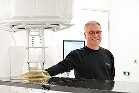 Ein lächelnder Mann steht an einem medizinischen Gerät in einem hellen Raum. Er trägt einen schwarzen Pullover und hat eine Brille auf. Hinter ihm befindet sich ein Monitor, der nicht näher zu erkennen ist.