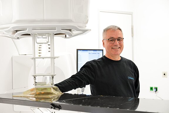 Ein lächelnder Mann steht an einem medizinischen Gerät in einem hellen Raum. Er trägt einen schwarzen Pullover und hat eine Brille auf. Hinter ihm befindet sich ein Monitor, der nicht näher zu erkennen ist.