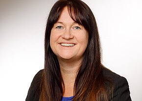 Porträt einer lächelnden Frau mit langen dunkelbraunen Haaren. Sie blickt in die Kamera, trägt ein dunkles Jackett und ein blaues Oberteil. Heller, neutraler Hintergrund; professionelles Business-Foto.