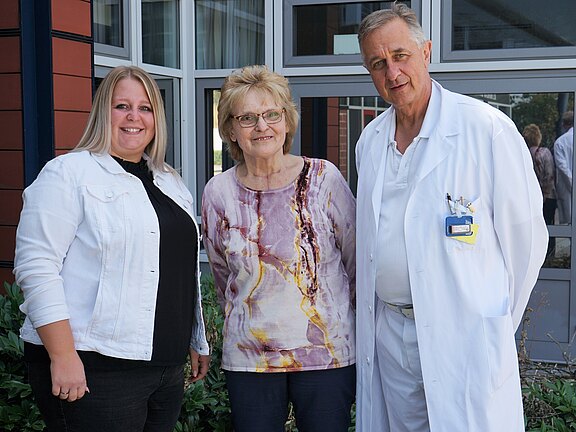 Drei Erwachsene posieren lächelnd vor einem Gebäude: links eine Frau in weißer Jacke, in der Mitte eine ältere Frau mit Brille, rechts ein Mann im weißen Arztkittel mit Ausweis. Sie stehen nah beieinander und wirken freundlich.