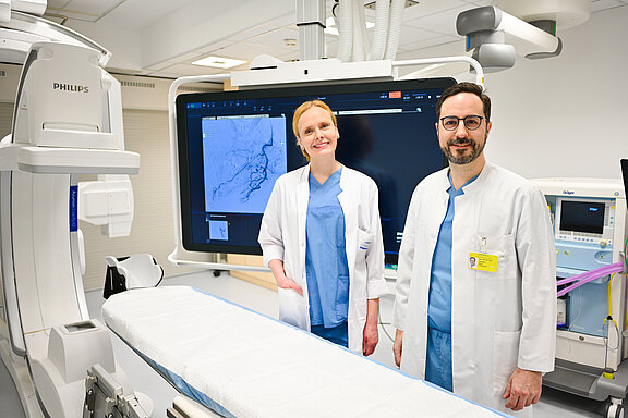 Zwei medizinische Fachkräfte in Kitteln stehen in einem modernen Untersuchungsraum. Hinter ihnen zeigt ein großer Monitor eine Angiografie- bzw. Röntgenaufnahme. Vorne steht ein steriler Untersuchungstisch. Die Szene vermittelt professionelle Vorbereitung für einen bildgesteuerten Eingriff.