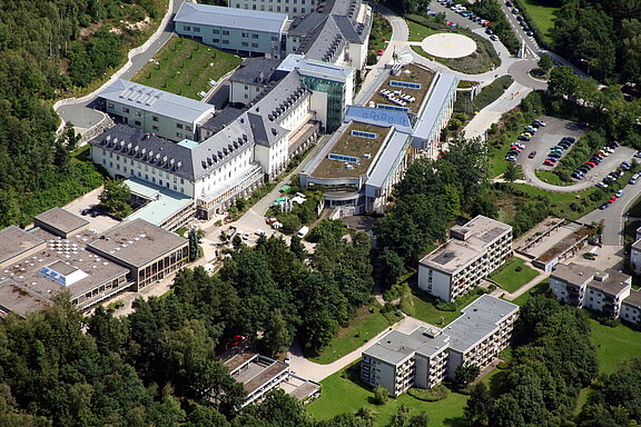 Luftaufnahme eines großen Klinikkomplexes in grüner Umgebung: mehrere verbundene Gebäude mit Flachdächern, teils begrünt; Wege und Parkplätze rundherum; rechts oben ein runder Hubschrauberlandeplatz. Wohn- oder Nebengebäude stehen nahe am Waldrand.