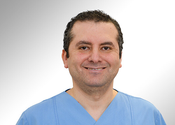 Porträt eines lächelnden Mannes in blauer medizinischer Kleidung vor neutralem Hintergrund, frontal zur Kamera.