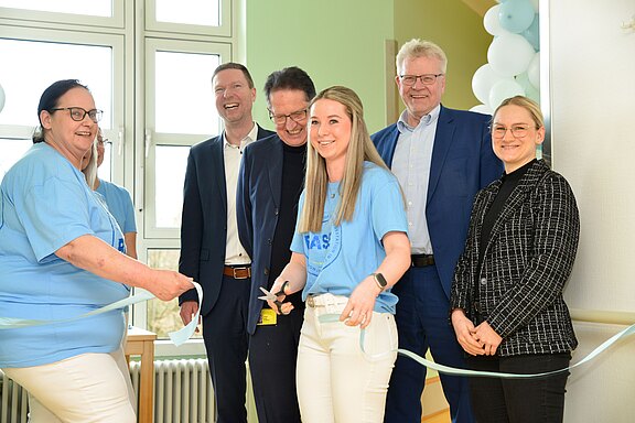 Gruppe lächelnder Erwachsener bei einer Eröffnungsfeier. Eine Frau in hellblauem T‑Shirt schneidet mit einer Schere ein blaues Band durch, während andere neben ihr stehen und lächeln. Ballons und Fenster im Hintergrund.
