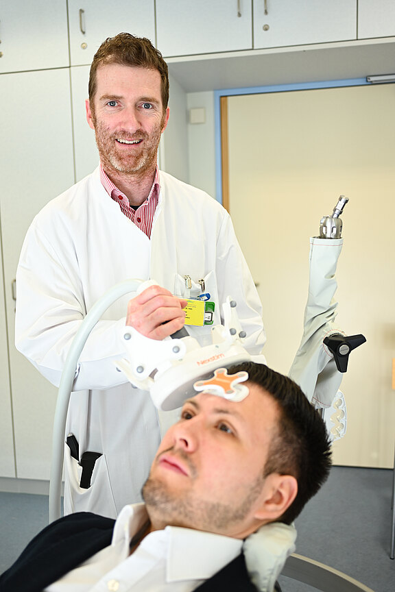 Ein Arzt in einem weißen Kittel hält ein medizinisches Gerät über dem Kopf eines Patienten, der in einem Behandlungsstuhl sitzt. Der Patient wirkt entspannt, während der Arzt sich mit ihm beschäftigt. Im Hintergrund sind medizinische Geräte sichtbar.