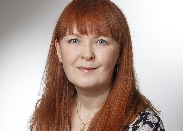 Porträt einer Frau mit langem roten Haar und Pony, leichtes Lächeln, Blick in die Kamera; goldene Ohrringe und Halskette; neutraler, heller Hintergrund.