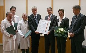 Gruppenfoto von fünf Personen in Anzug, die eine Urkunde halten, umgeben von medizinischem Personal in Kitteln. Sie stehen in einem Raum mit hellen Wänden, einige Personen halten Dokumente und eine Blumenstrauß wird überreicht. Die Stimmung ist feierlich und anerkennend.