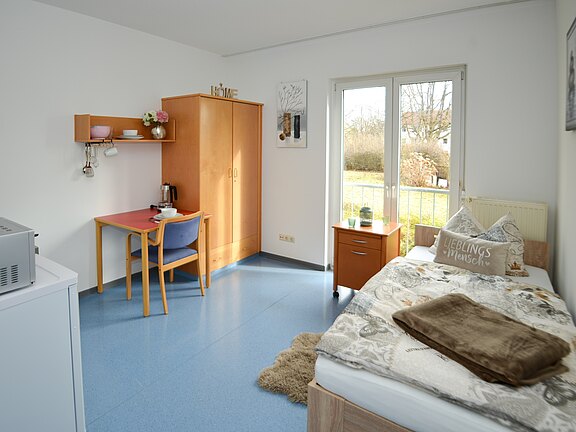 Helles, aufgeräumtes Einzelzimmer: rechts ein Einzelbett mit Kissen und Decke, daneben ein kleiner Nachttisch. Links ein Schreibtisch mit Stuhl, darüber Regal; daneben Kleiderschrank. Große Terrassentür mit Blick ins Grüne; schlichte, funktionale Einrichtung.