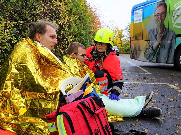 Drei Personen sitzen am Straßenrand, eingewickelt in goldene Rettungsdecken. Eine Person in Rettungsuniform hilft den anderen, die sichtbar verunsichert sind. Im Hintergrund ist ein Bus mit einem Werbebild zu sehen. Die Szene vermittelt einen ernsten Erste-Hilfe-Einsatz.