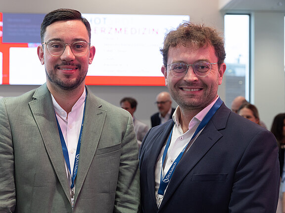 Zwei Männer in Anzügen mit blauen Konferenzlanyards stehen nebeneinander und lächeln in die Kamera. Hinter ihnen ein heller Messestand mit unscharfem Bildschirm und mehreren Personen im Hintergrund. Fokus auf freundliches Porträt während einer Fachveranstaltung.
