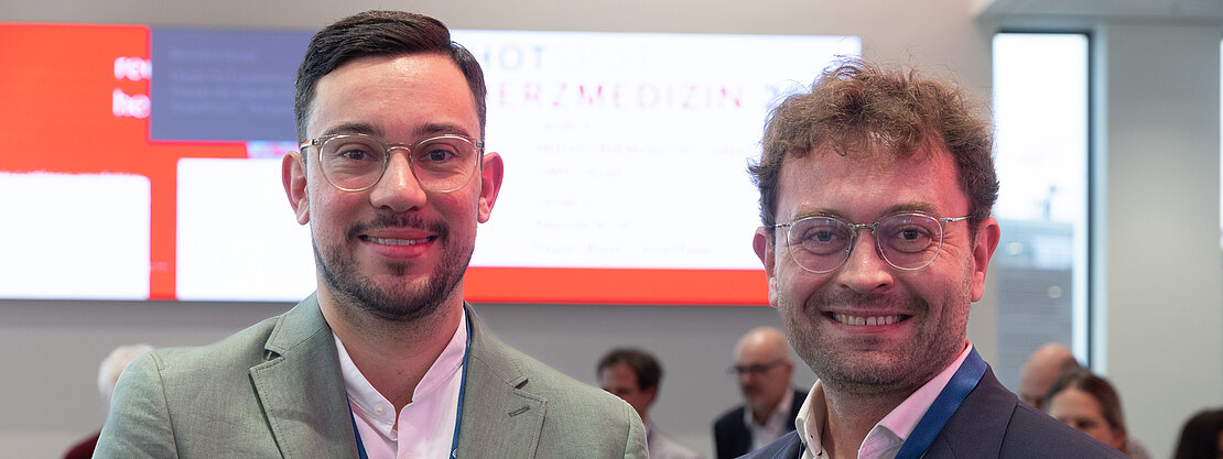 Zwei Männer in Anzügen mit blauen Konferenzlanyards stehen nebeneinander und lächeln in die Kamera. Hinter ihnen ein heller Messestand mit unscharfem Bildschirm und mehreren Personen im Hintergrund. Fokus auf freundliches Porträt während einer Fachveranstaltung.