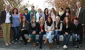 Eine Gruppe von 17 Personen posiert gemeinsam im Freien. Die Mehrheit sind junge Frauen, die lächeln und in modischer Kleidung gekleidet sind. Im Hintergrund sind Bäume mit herbstlichen Blättern sichtbar. Einige der Anwesenden tragen Schals. Ein Mann steht am rechten Rand der Gruppe.