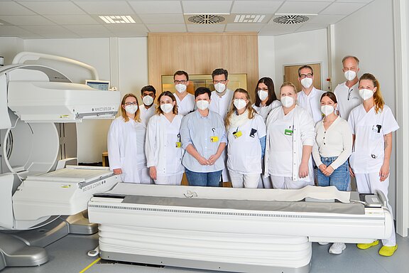 Gruppe von medizinischem Fachpersonal in weißen Kitteln und Masken steht in einem Klinikraum neben einem großen bildgebenden Gerät; sie posieren für ein Teamfoto.