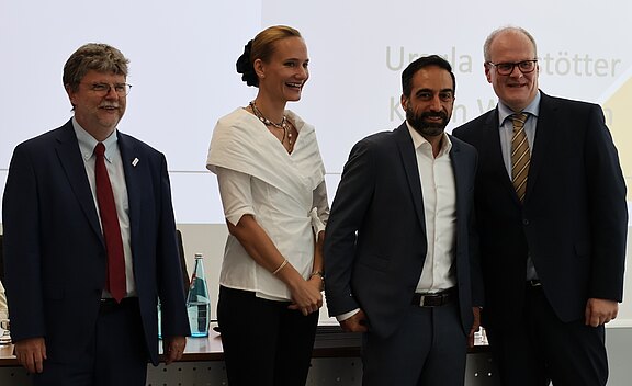 Vier Personen stehen nebeneinander in einem Konferenzraum und posieren lächelnd für ein Foto. Zwei Männer im Anzug, eine Frau in weißer Bluse und ein Mann im dunklen Anzug. Im Hintergrund sind eine Präsentationsfolie und sitzende Teilnehmende zu sehen.