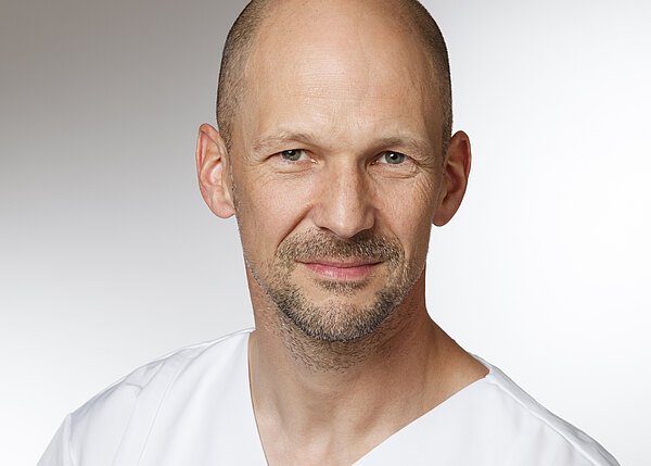 Porträt eines lächelnden, glatzköpfigen Mannes in weißer medizinischer Kleidung vor neutralem Hintergrund. Nahaufnahme bis zur Brust, ruhiger, freundlicher Gesichtsausdruck.