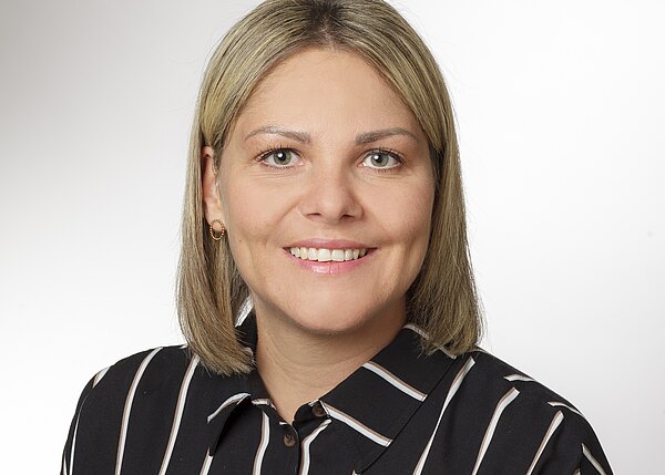 Porträtfoto einer lächelnden erwachsenen Frau mit schulterlangen blonden Haaren, vor neutralem Hintergrund. Sie trägt eine schwarz-weiße, vertikal gestreifte Bluse und kleine goldene Ohrringe, blickt direkt in die Kamera.