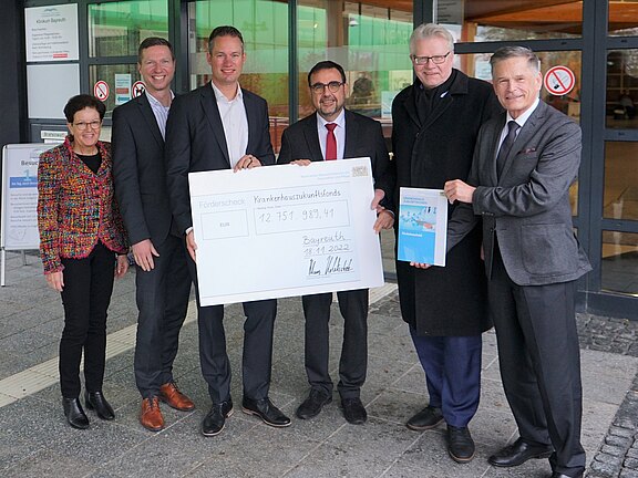 Sechs Personen vor einem Gebäude präsentieren einen übergroßen Scheck des Krankenhausstrukturfonds; feierliche Übergabe von Fördermitteln. Eine Person zeigt zusätzlich eine Broschüre. Alle lächeln in die Kamera.