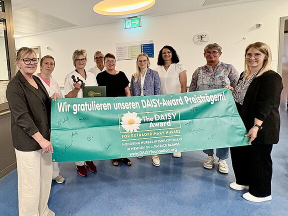Gruppe von Pflegekräften in einem Krankenhausflur hält ein großes türkisfarbenes Banner mit dem Text: Wir gratulieren unseren DAISY-Award Preisträgern! The DAISY Award – For Extraordinary Nurses. Feierliche Auszeichnung, alle lächeln in die Kamera.