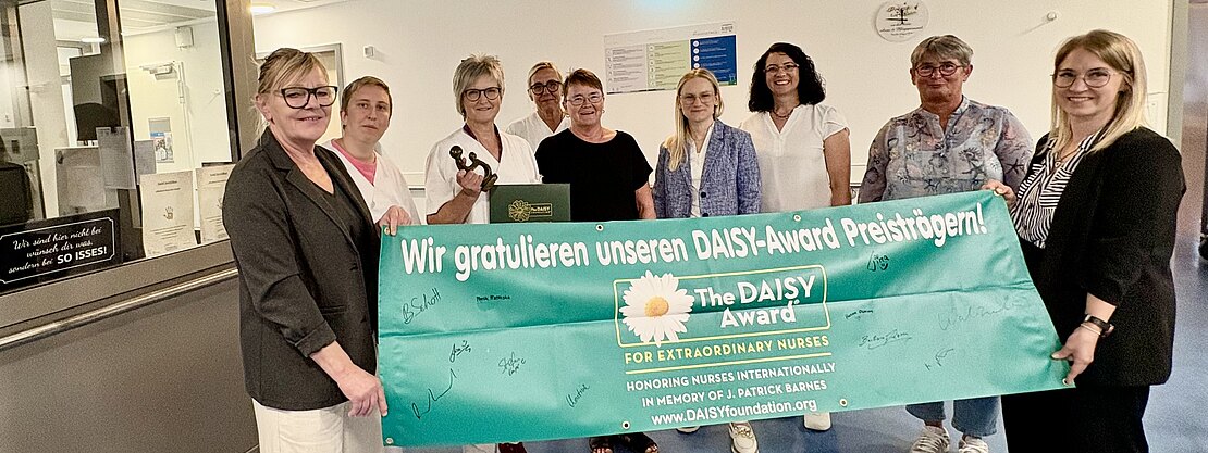Gruppe von Pflegekräften in einem Krankenhausflur hält ein großes türkisfarbenes Banner mit dem Text: Wir gratulieren unseren DAISY-Award Preisträgern! The DAISY Award – For Extraordinary Nurses. Feierliche Auszeichnung, alle lächeln in die Kamera.
