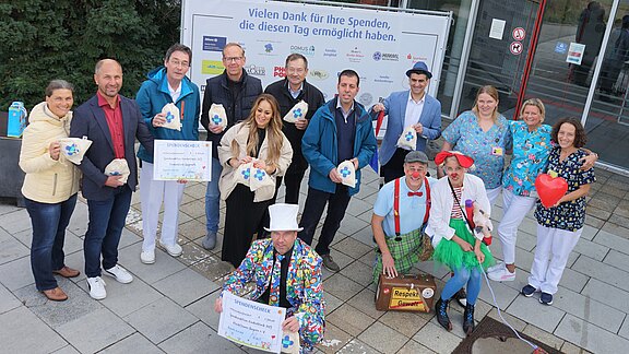 Gruppenfoto vor einem Gebäude: Krankenhausmitarbeitende und zwei Klinikclowns halten Spendenherzen und einen großen Scheck. Hinter ihnen ein Banner mit Dank an Spender. Stimmung fröhlich; Aktion offenbar Benefiz oder Spendenübergabe.