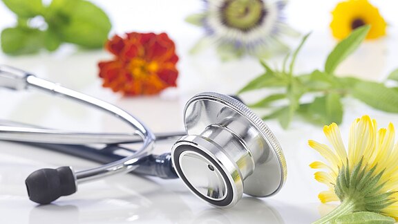 Ein silbernes Stethoskop liegt auf einer weißen Fläche, umgeben von bunten Heilblüten und Kräutern – Symbol für die Verbindung von Medizin und Naturheilkunde.