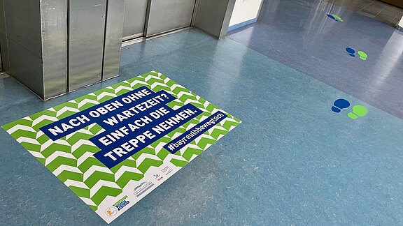 Am Aufzug klebt ein großes Bodenplakat mit grünen Zickzackstreifen. Darauf steht: „Nach oben ohne Wartezeit? Einfach die Treppe nehmen.“ Daneben farbige Fußspuren weisen den Weg. Botschaft: Statt auf den Aufzug zu warten, lieber die Treppe benutzen.