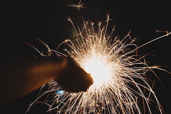 Dunkler Hintergrund; eine Hand hält eine leuchtende Wunderkerze. Funken sprühen in alle Richtungen und bilden helle, goldene Linien und Sterne. Der Fokus liegt auf dem intensiven Licht und den bewegten Funken.