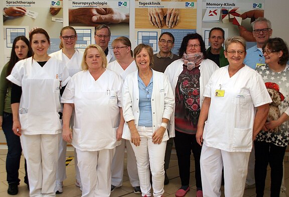 Gruppe von Klinikmitarbeitenden posiert in einem Flur; mehrere Personen tragen weiße Pflege- oder Arztkleidung und Namensschilder. Hinter ihnen hängen Poster zu Sprachtherapie, Neuropsychologie, Tagesklinik und Memory-Klinik. Atmosphäre: Teamfoto im Gesundheitszentrum.