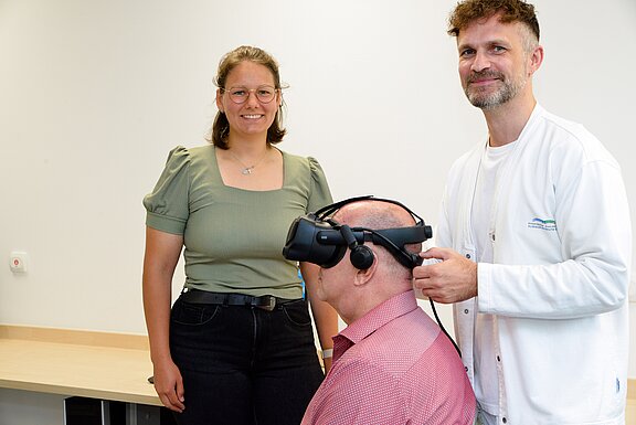 Drei Personen stehen in einem modernen Raum. Eine Frau mit Brille lächelt in die Kamera, während ein Mann einer anderen Person, die ein Virtual-Reality-Headset trägt, bei der Einstellung hilft. Der Fokus liegt auf der Interaktion zwischen ihnen und der Nutzung der VR-Technologie.