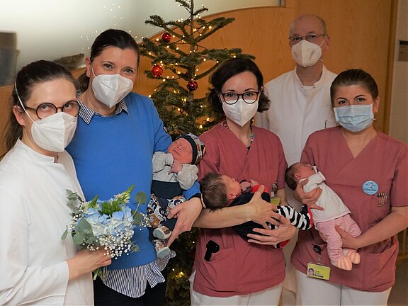 Gruppe von fünf Erwachsenen, überwiegend in Klinikbekleidung, posiert mit drei Neugeborenen. Alle tragen Schutzmasken. Eine Person hält einen Blumenstrauß. Im Hintergrund steht ein geschmückter Weihnachtsbaum. Szene wirkt wie ein Krankenhausfoto zur Geburt.