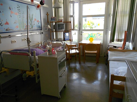 Zweibettiges Krankenhauszimmer: links ein verstellbares Patientenbett mit Nachttisch und Infusionsständer, rechts ein weiteres Bett. Am Fenster ein kleiner Tisch mit zwei Stühlen, Tageslicht fällt herein. Schlichter, sauberer Raum für Patientenaufenthalt.