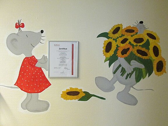 Wandbild mit zwei Cartoon-Mäusen: links eine graue Maus im roten Kleid lächelt, rechts bringt eine andere Maus einen großen Strauß Sonnenblumen. Eine einzelne Sonnenblume liegt am Boden. Fröhliche, kindliche Szene, als würde die Maus ein Geschenk erhalten.