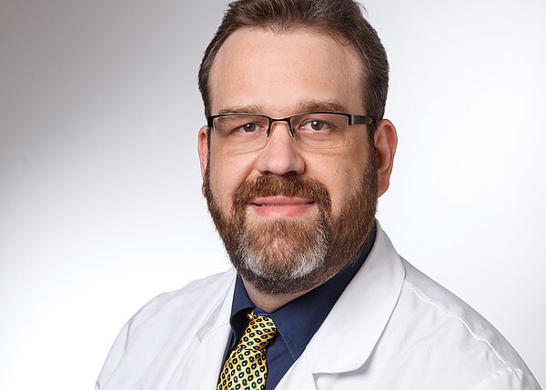 Porträt eines Mannes mit Brille und Vollbart, der einen weißen Kittel trägt. Er lächelt freundlich in die Kamera, vor einem hellen, neutralen Hintergrund. Der Kittel deutet darauf hin, dass er im medizinischen Bereich tätig ist.