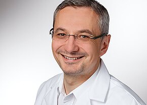 Dr. med. Marek Pizon Porträt einer lächelnden Person mit Brille und kurzem Haar, die einen weißen Kittel trägt; neutraler, heller Hintergrund.