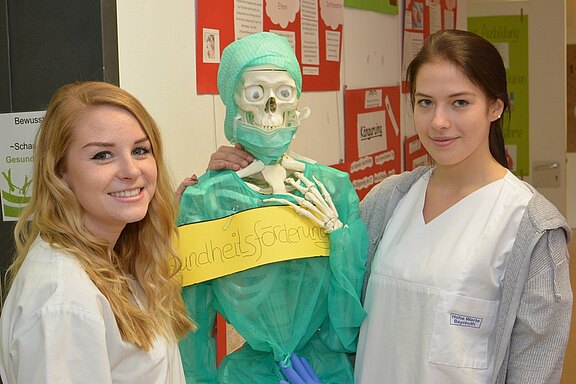 Zwei Frauen in medizinischer Kleidung stehen nebeneinander und lächeln. Zwischen ihnen steht ein Skelett, das mit grüner Schutzkleidung und einem Schild mit der Aufschrift "Gesundheitsförderung" ausgestattet ist. Im Hintergrund sind mehrere Plakate zu erkennen.