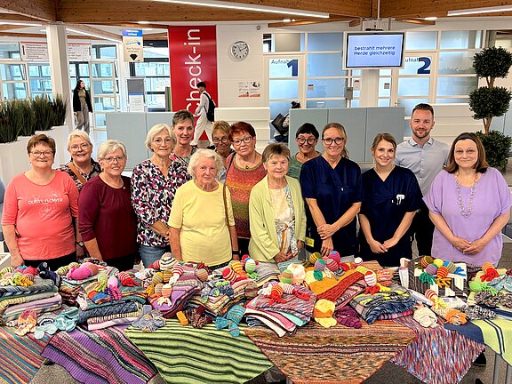 Gruppe lächelnder Menschen in einem hellen Klinik-Wartebereich steht hinter Tischen voller bunter, handgestrickter Socken, Mützen und Decken. Die Präsentation wirkt wie eine Spenden- oder Gemeinschaftsaktion; einige tragen Berufsbekleidung, andere Freizeitkleidung.