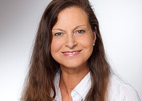 Porträtfoto: Eine lächelnde erwachsene Frau mit langen braunen Haaren und Perlenohrringen blickt in die Kamera. Sie trägt eine weiße Bluse. Neutraler, heller Hintergrund.