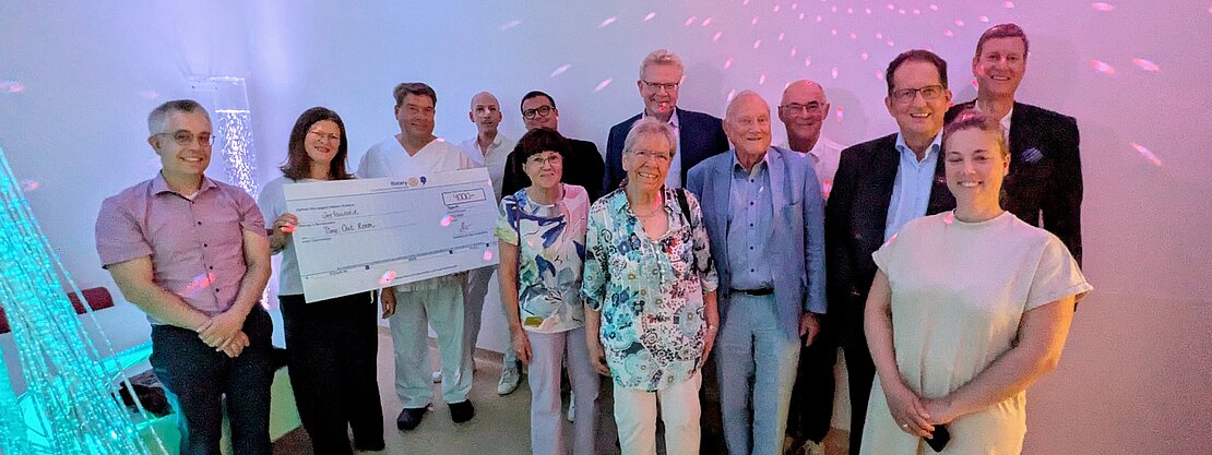 Eine Gruppe von Personen steht in einem hellen Raum, in dem ein großer Scheck präsentiert wird. Die Menschen lächeln und posieren zusammen. Im Hintergrund sind farbige Lichter und eine dekorative Wandgestaltung zu sehen. Der Scheck symbolisiert eine Spende für einen guten Zweck.