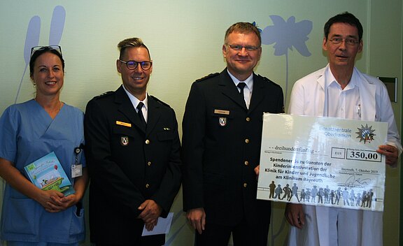 Zwei Polizisten, eine Pflegekraft und ein Arzt präsentieren einen großen Spenden-Scheck. Betrag: 350 Euro für die Kinder- und Jugendklinik. Die Gruppe steht zusammen im Krankenhaus und lächelt in die Kamera.