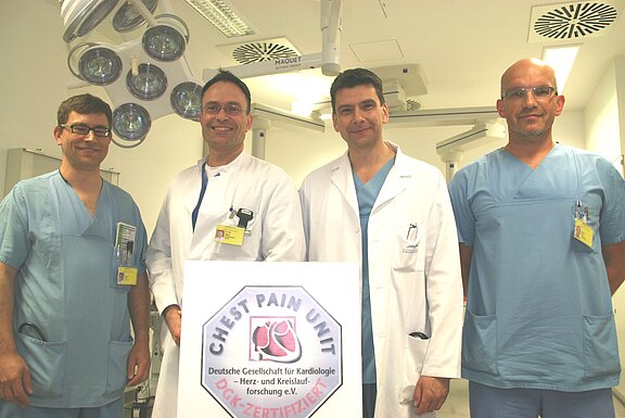 Vier medizinische Fachkräfte in einem Untersuchungsraum stehen nebeneinander und lächeln. Vor ihnen ein Schild mit der Aufschrift „Chest Pain Unit“ und Hinweis auf die Zertifizierung durch die Deutsche Gesellschaft für Kardiologie. Aussage: Team präsentiert eine zertifizierte Einheit zur Behandlung von Brustschmerzen.