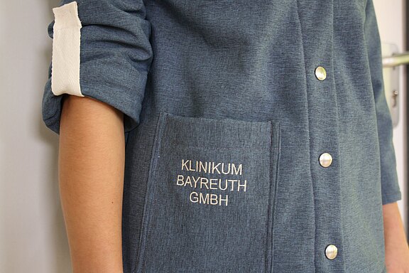 Nahaufnahme einer Person in blauer Klinikjacke mit aufgerolltem Ärmel. Auf der Brusttasche steht in Weiß „Klinikum Bayreuth GmbH“; daneben sind silberne Knöpfe sichtbar.